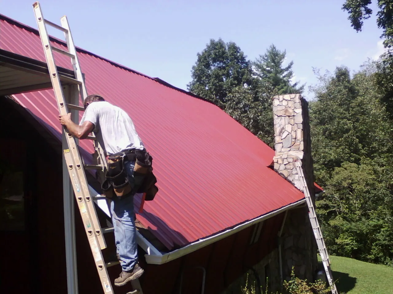 Roofer on a ladder beside a red metal roof and stone chimney — Soffit & Fascia Repair by Roofing Brothers in Carnesville